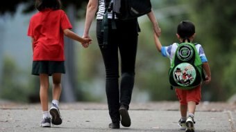 A mother walks holding the hands of two children on their way to school holding backpacks.