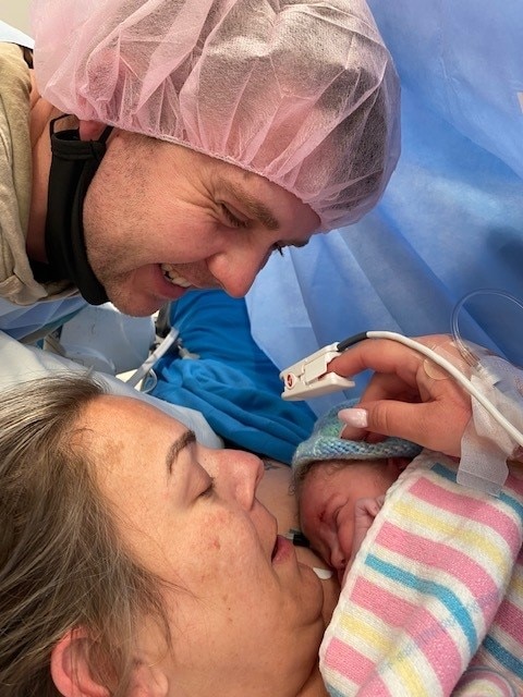 A woman holding her newborn baby in a hospital bed, as her partner looks on smiling.