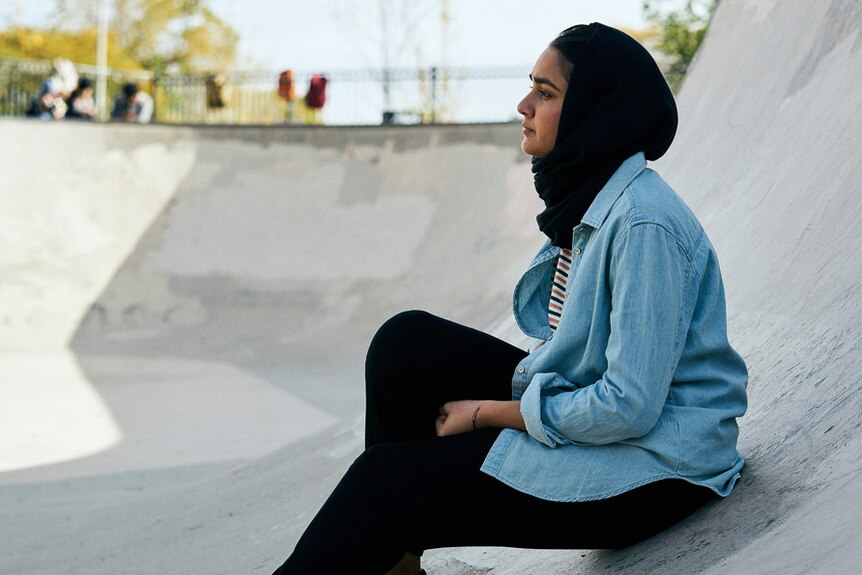 Girl in headscarf sits in cement skate bowl outdoors, daylight, wearing blue button down shirt and black pants.