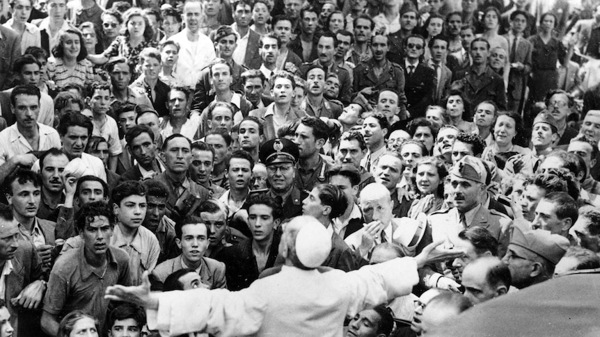 Men, women and soldiers gather around Pope Pius XII, his arms outstretched.