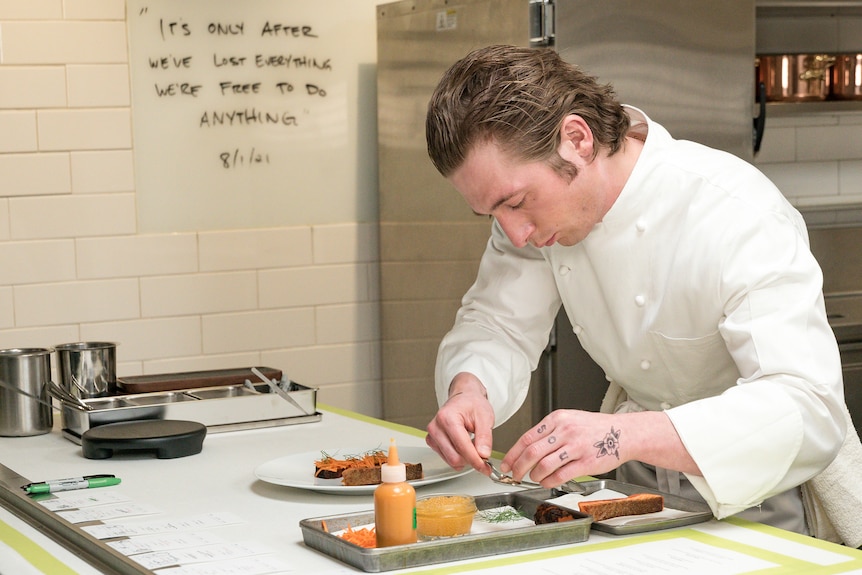 A young white man in a white chef shirt and hair slicked back, carefully prepares an elaborate dish in a commercial kitchen