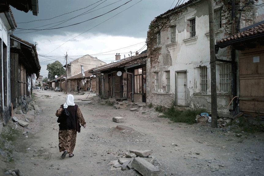 A woman walks down a dirt street edged with houses in disrepair.