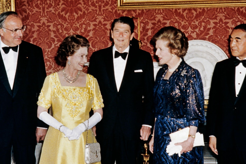 Queen Elizabeth II and Margaret Thatcher face each other in a room full of men.