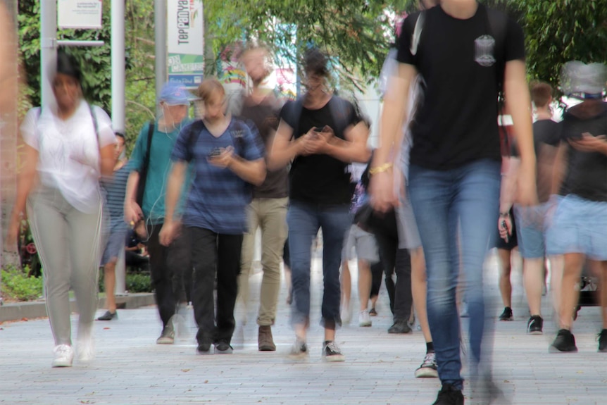 Студенти се разхождат през кампуса, някои гледат телефоните си.  Снимката е размазана, което прави лицата неразпознаваеми.