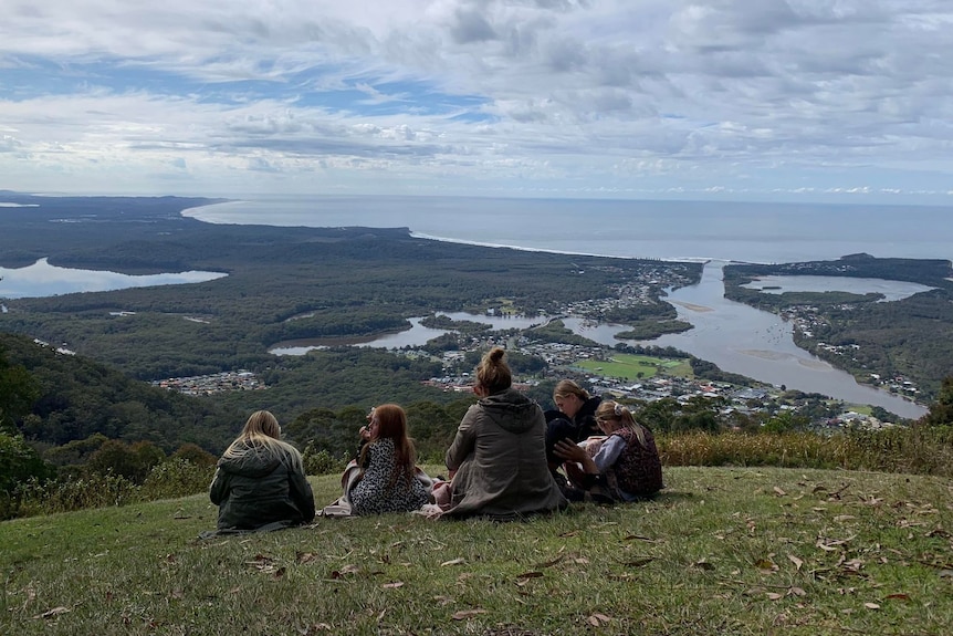 A mother and four kids sit on a hill with a picnic and look at the view of a coastal town