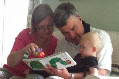 An infant sits on the lap of a man and woman as they read him The Very Hungry Caterpillar.