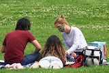 Two women and a man sitting on a rug on green grass in a park.