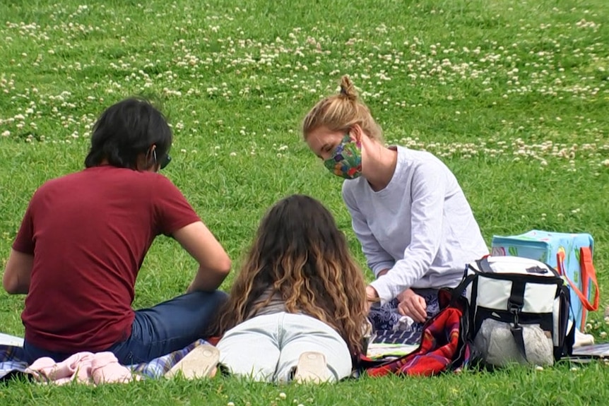 Two women and a man sitting on a rug on green grass in a park.