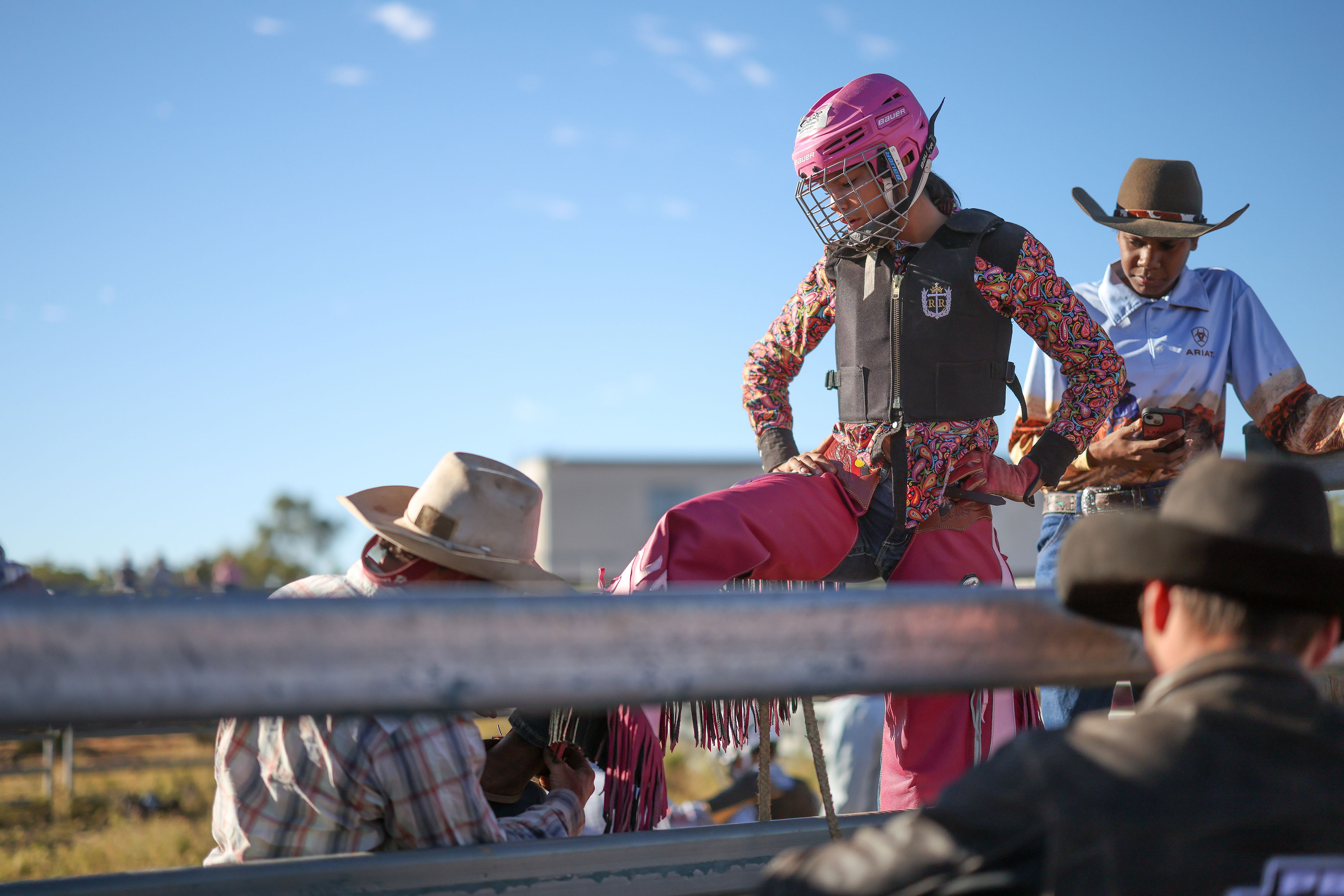 Lareece Daley-Kennedy stands in a pink helmet and protective vest beside the rodeo chute as adults prepare her for a ride.