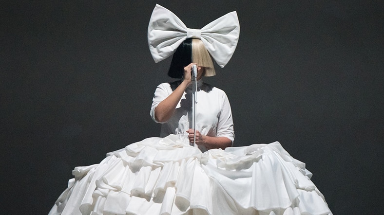 Colour photo of Sia performing on stage wearing large bow, dress and wig in 2016.