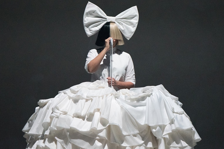 Colour photo of Sia performing on stage wearing large bow, dress and wig in 2016.