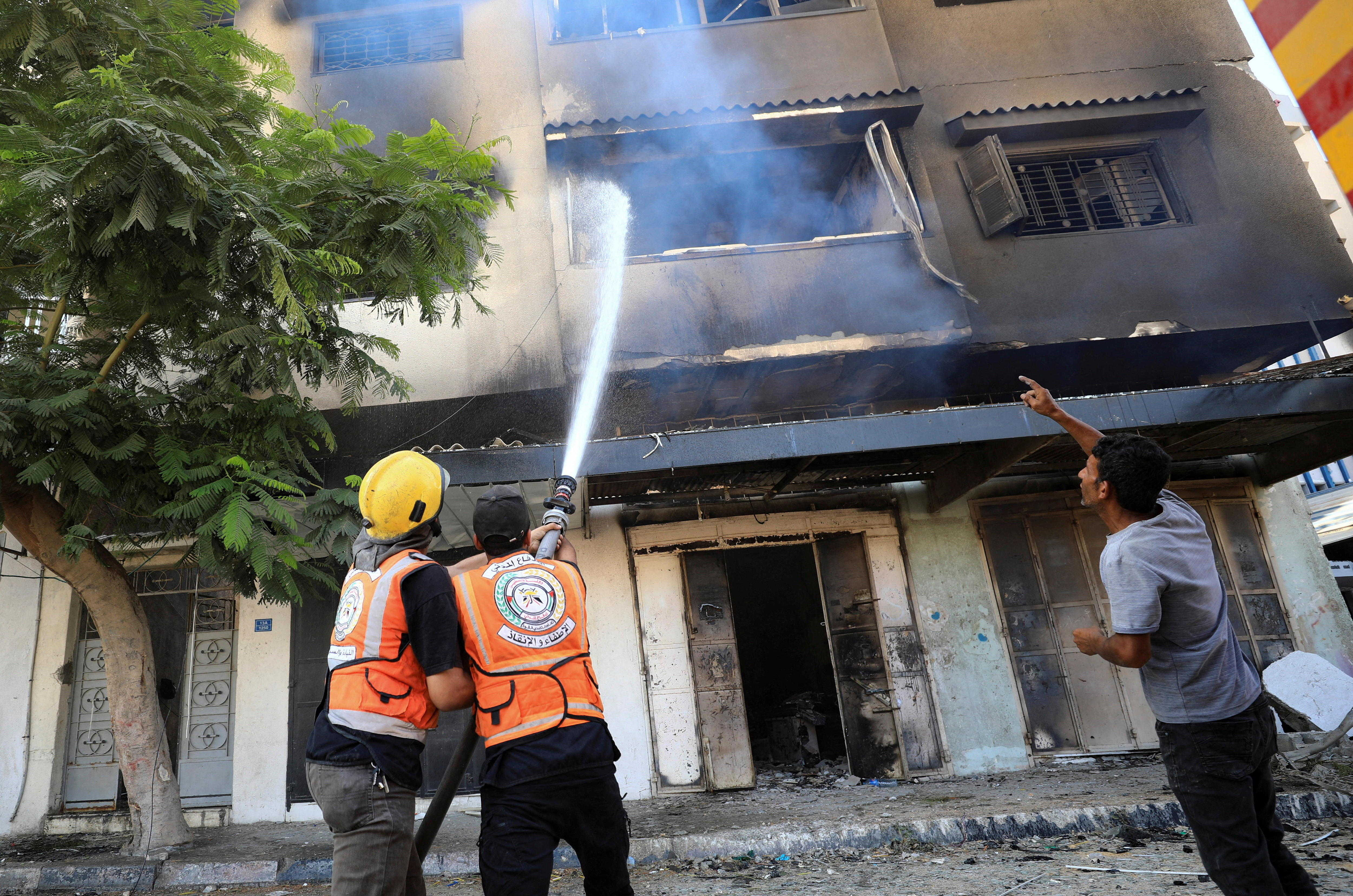 Two firefighters spray water into a window, while a man in a grey shirt points up at it.