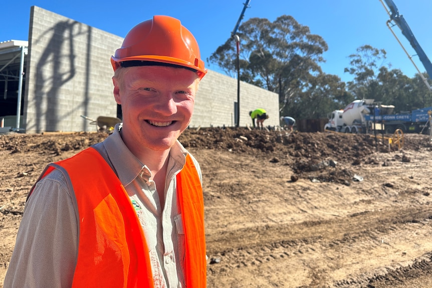 A smiling Caucasian man high-viz orange hat and vest, in front of a construction site, dirt ground, sun on his face, blue sky.