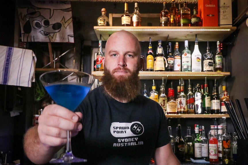 A man holds out a blue cocktail, standing behind a bar with rows of alcohol bottles on shelves behind him.
