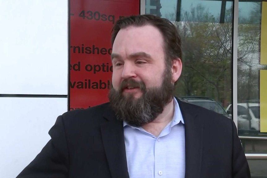 A bearded, dark-haired man in a suit stands outside a building.