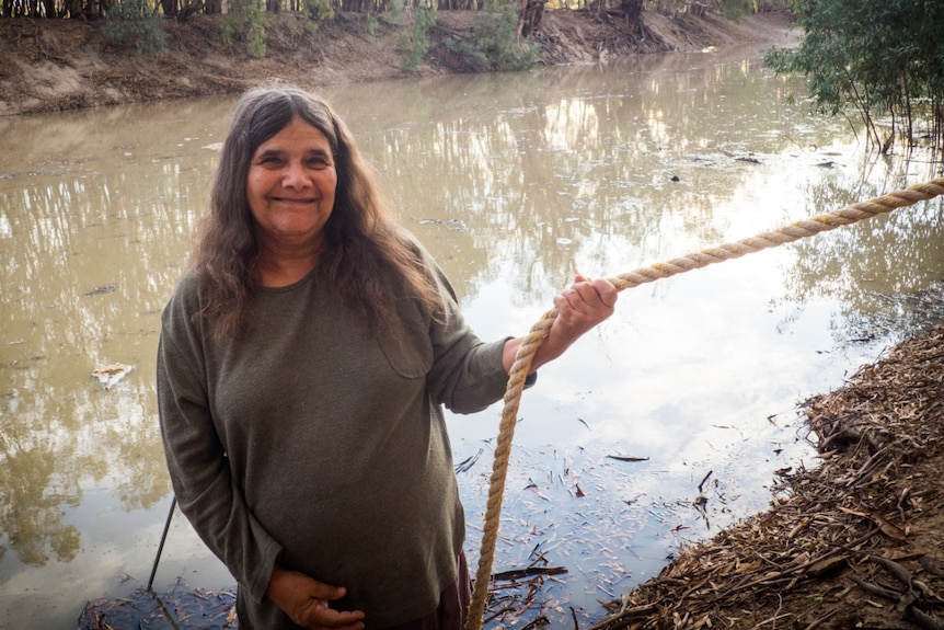 Paakantyi woman Patricia Johnson smiling and holding a rope for balance as the Darling flows behind her.