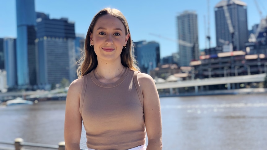 A young woman looks to the camera and slightly smiles while standing in front of the Brisbane CBD from across a river.