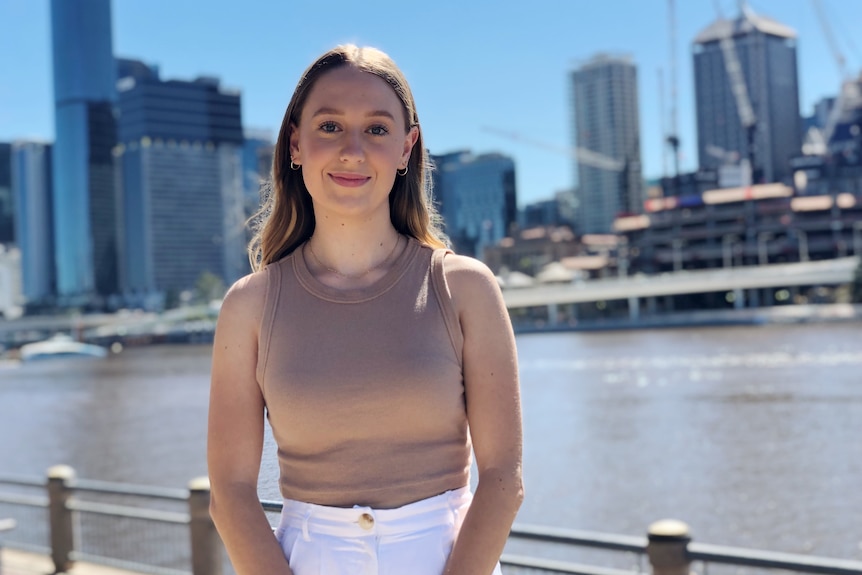 A young woman looks to the camera and slightly smiles while standing in front of the Brisbane CBD from across a river.