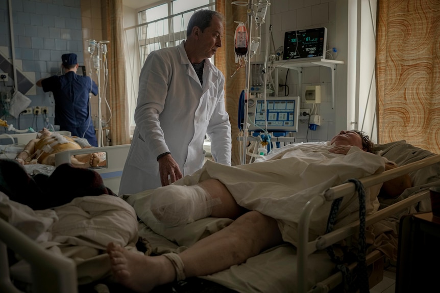 A male doctor speaks to a woman lying in a hospital bed. Her right leg has been amputated and bandaged.