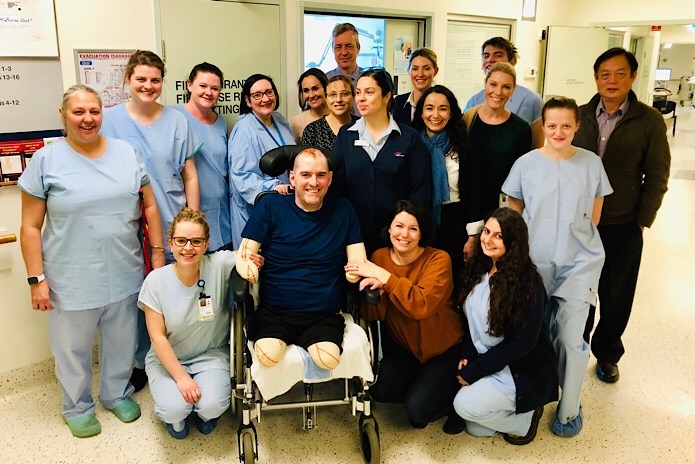 A group photo of medical staff in scrubs with a man with no limbs in a chair, smiling