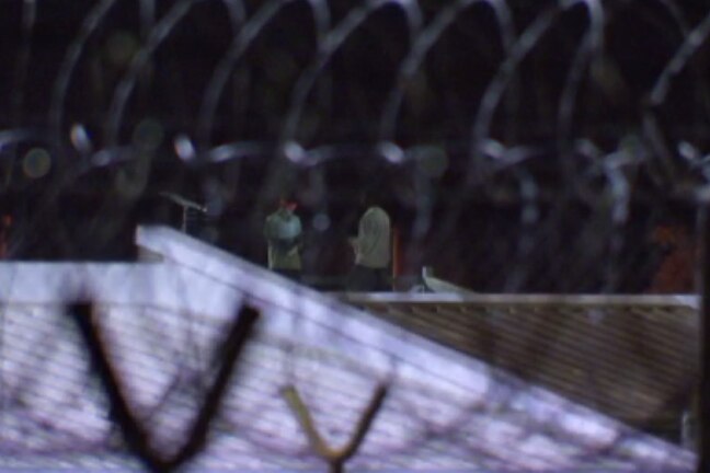 Blurry image of two boys on a roof of a building at night, seen through barbed wire fence