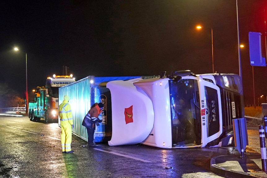 A truck lies on its side with two people standing next to it