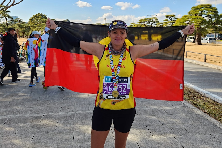 A woman smiles in running gear while holding up an Australian Aboriginal flag.