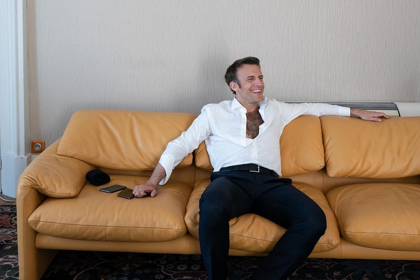 Emmanuel Macron sits on a couch with part of his white shirt unbuttoned.