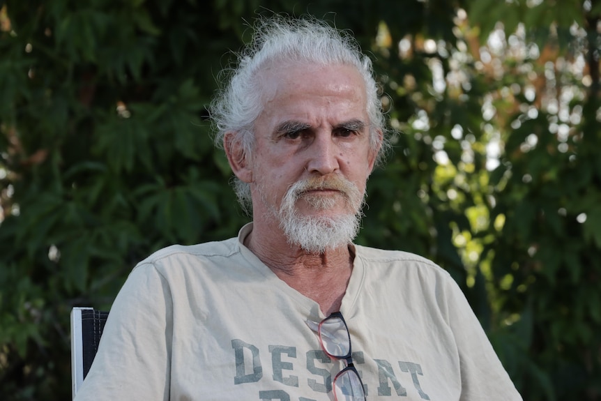 A man with gray hair and beard looking at the camera, sitting on a chair in a garden.