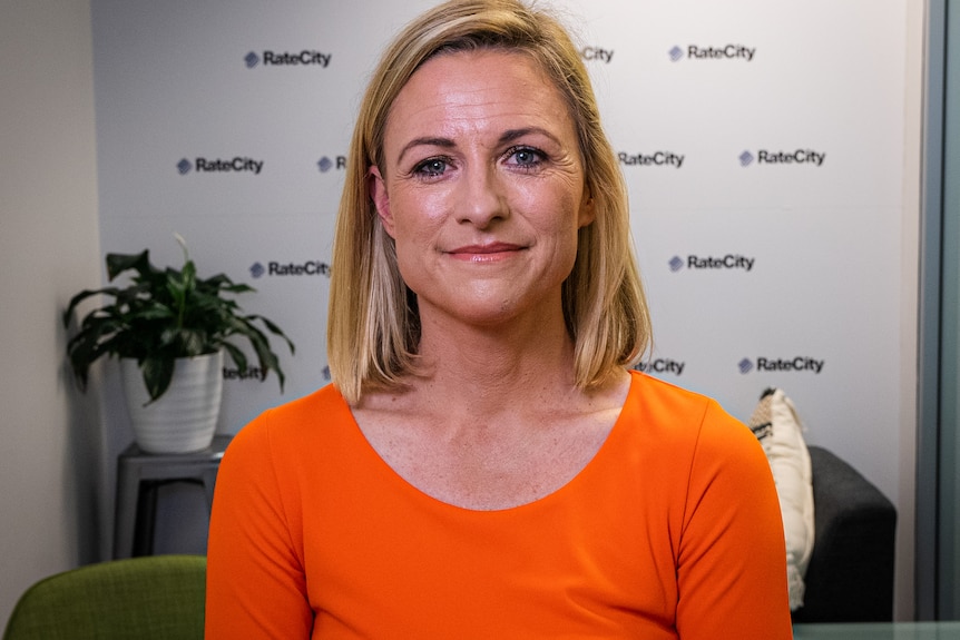 A blonde woman with an orange top smiles and looks directly at the camera.