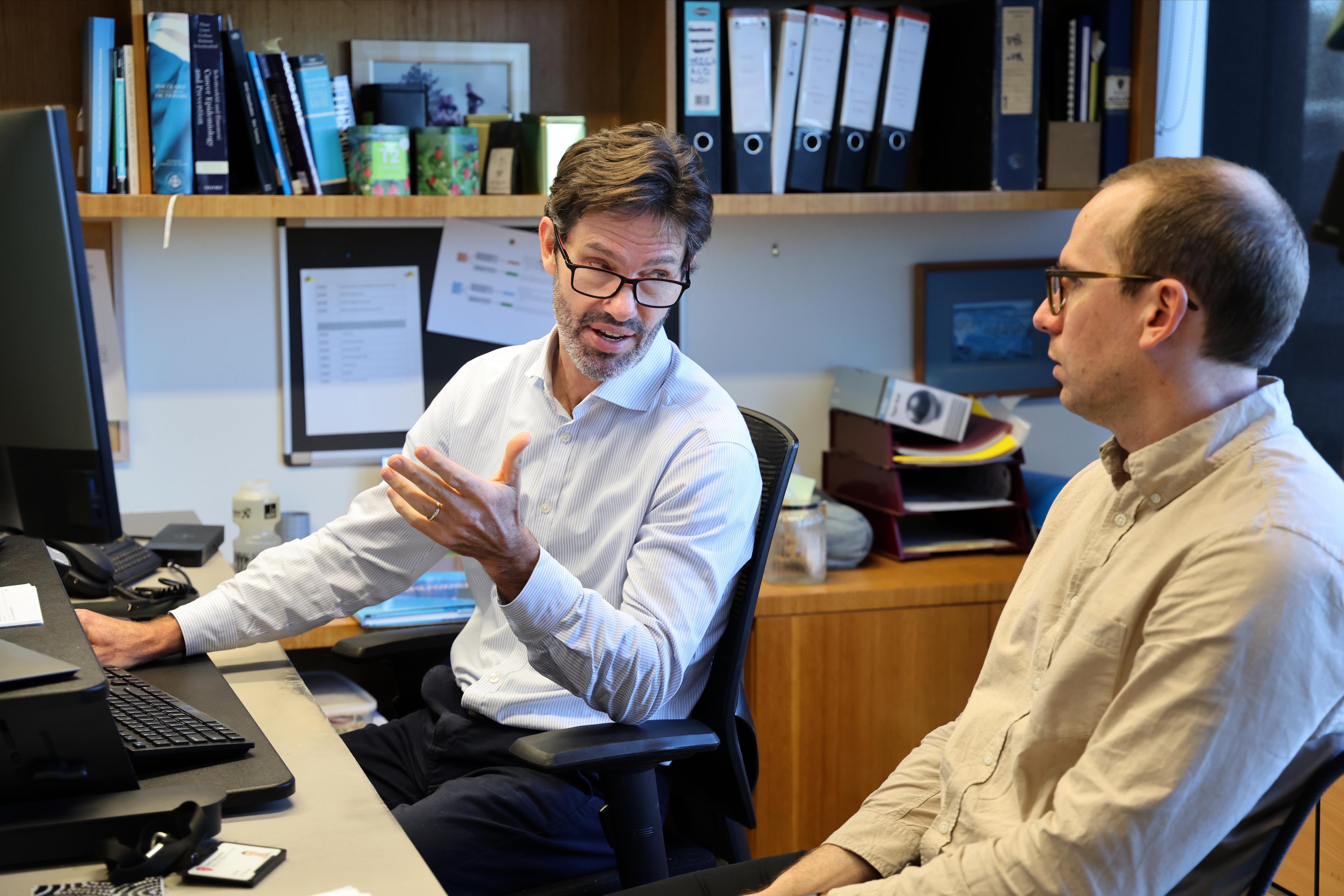 Two men sit at a desk in front of a computer in an office.