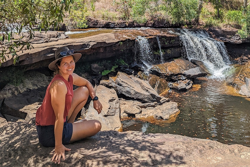 A woman wearing an orange top, hat and shorts sits beside a waterfall
