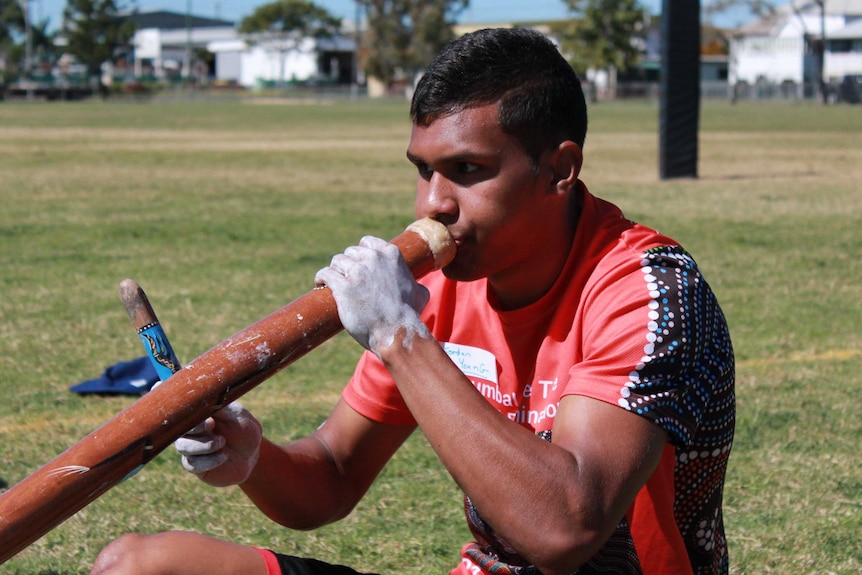 Young Indigenous man seated on ground playing the didgeridoo