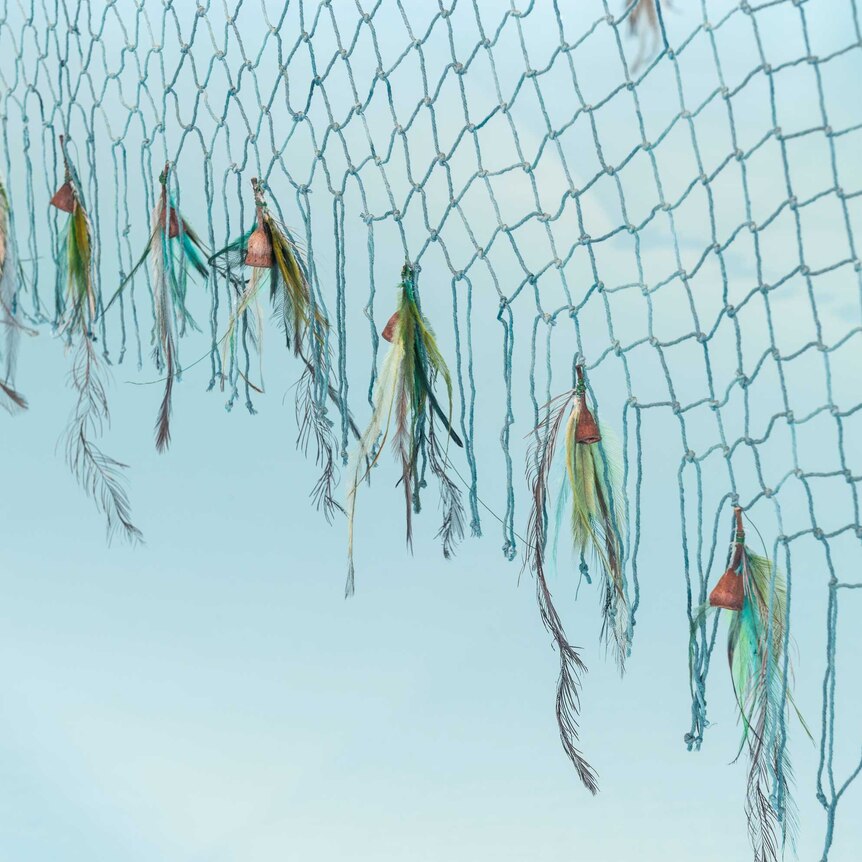You view a closeup of a yarn net against a baby blue backdrop, with gumnuts and bird feathers attached to the net's ends.