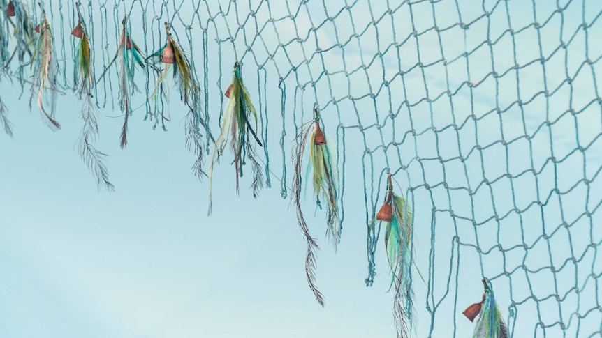 You view a closeup of a yarn net against a baby blue backdrop, with gumnuts and bird feathers attached to the net's ends.