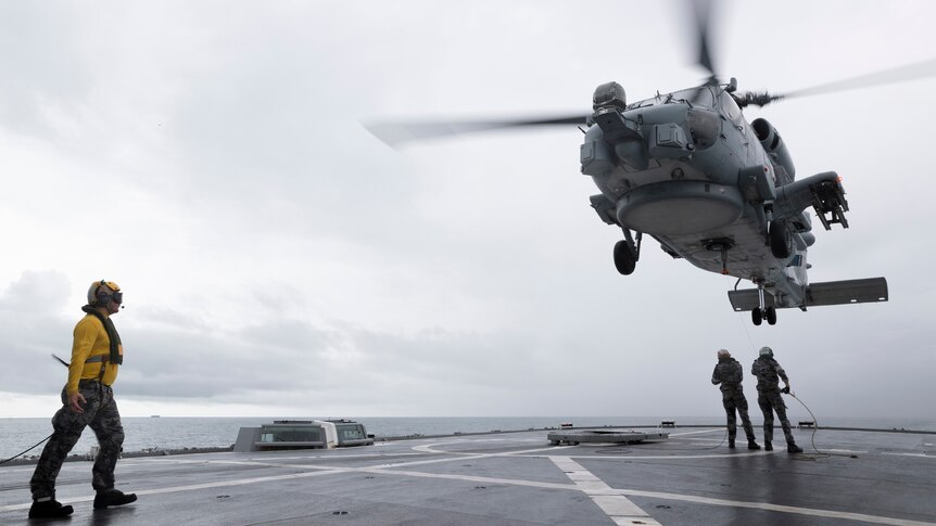 A helicopter lands aboard a ship under a grey sky, assisted by three sailors.