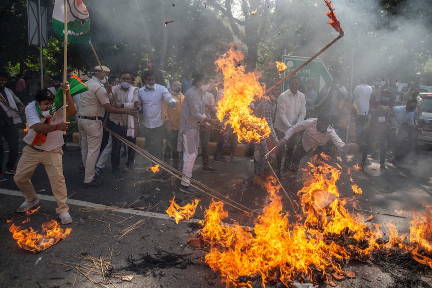 A number of Indian men stand around a large street fire, in which is burning effigies and flags