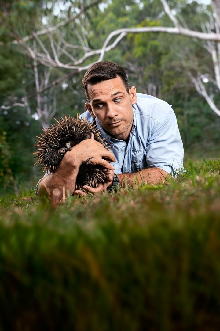 A man in a blue shirt lying on grass closely observing an echidna he's gently holding, with trees in the background.