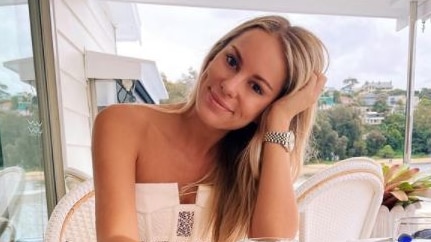 A young blonde woman sitting at a table on a veranda, smiling.