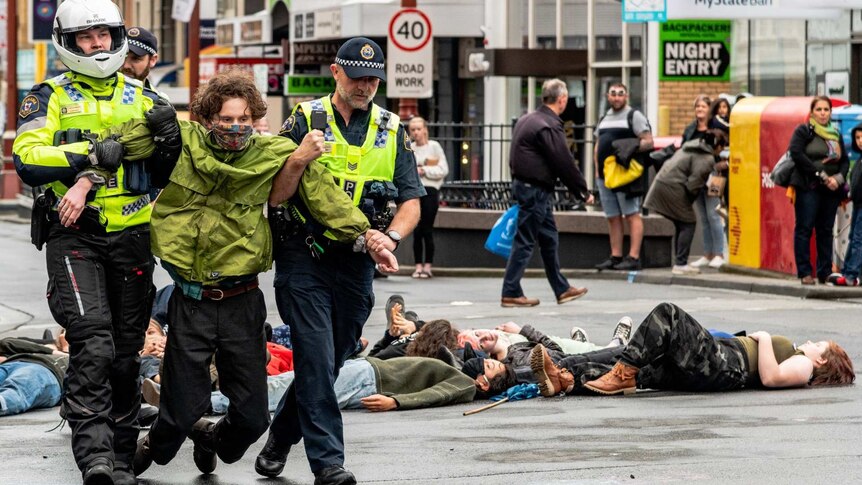 a protester wearing a face mask is dragged away by two police officers as other protesters lay on the ground behind them