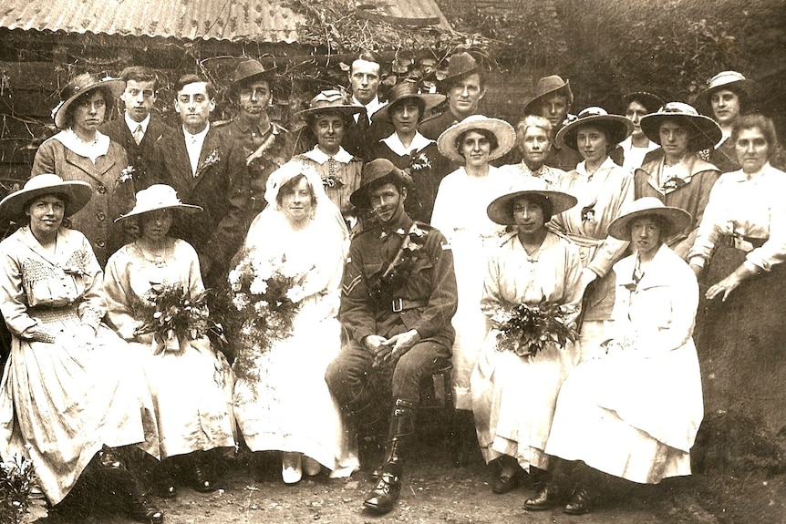 An old sepia wedding photo featuring a bride and groom in uniform surrounded by women in dresses and men in suits.