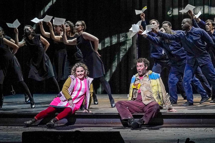 A white female opera singer sits onstage, wearing a pink dress, beside a white male opera singer in painting clothes, singing.