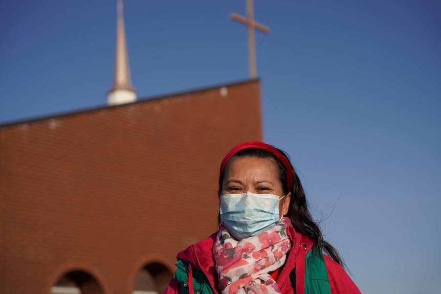 woman in pink with a blue face mask looks at the camera infront of a building with a cross on it