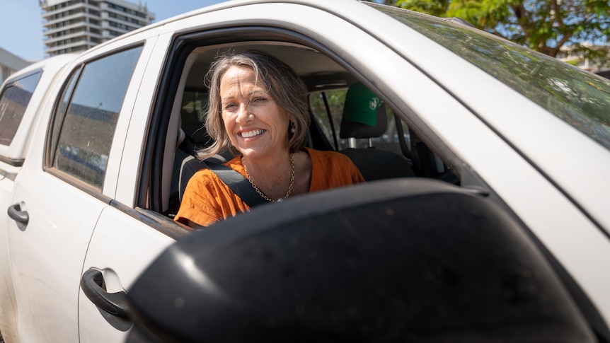 Photo of a woman with a light tan and cropped hair in a driver's seat. She is wearing an orange shirt.