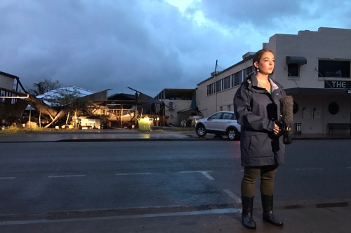 Allyson Horn stands outside a damaged building, holds a microphone doing an early morning cross from Proserpine.