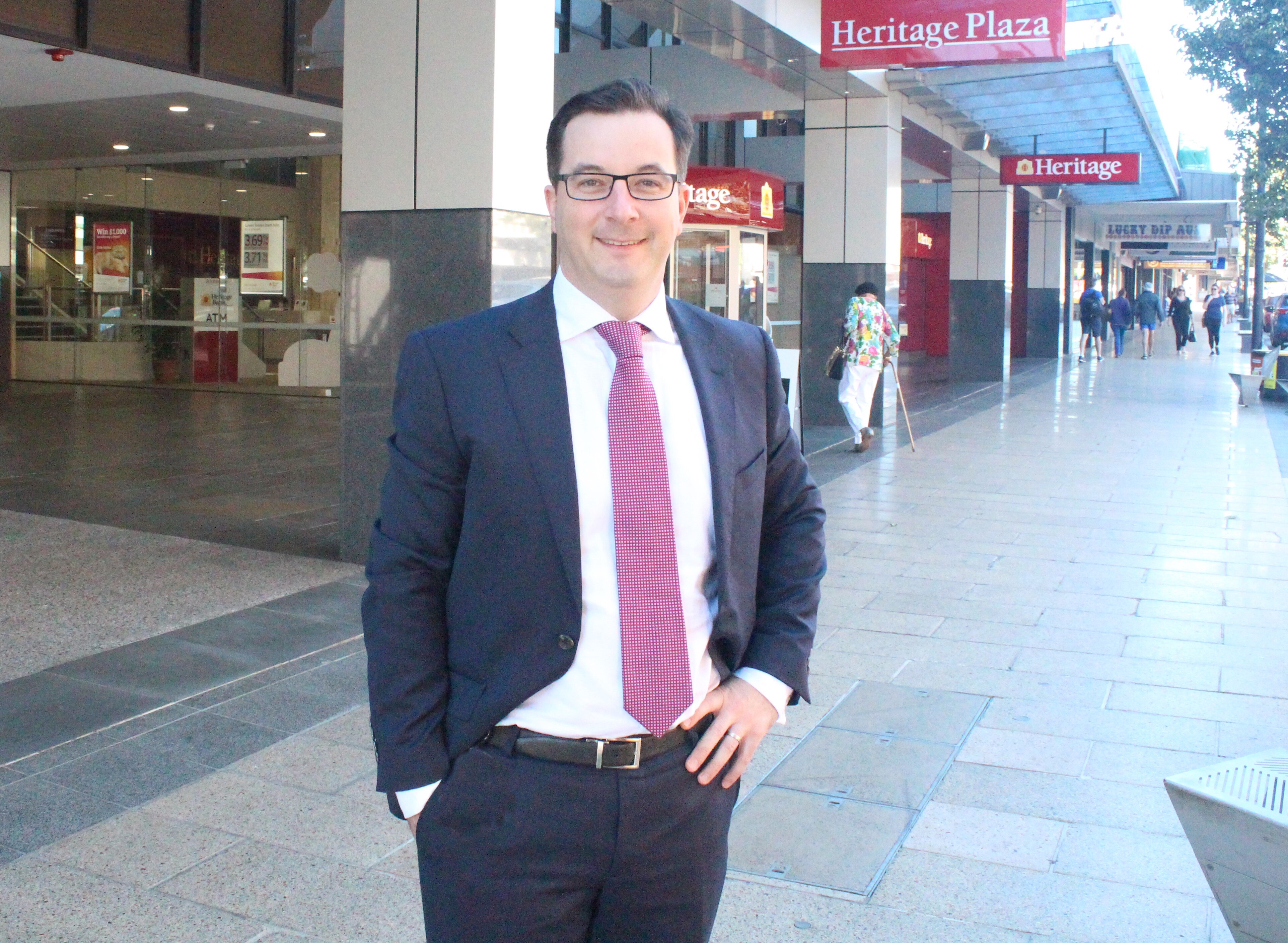A middle-aged smiling man with glasses wearing a suit on the street, pink tie.