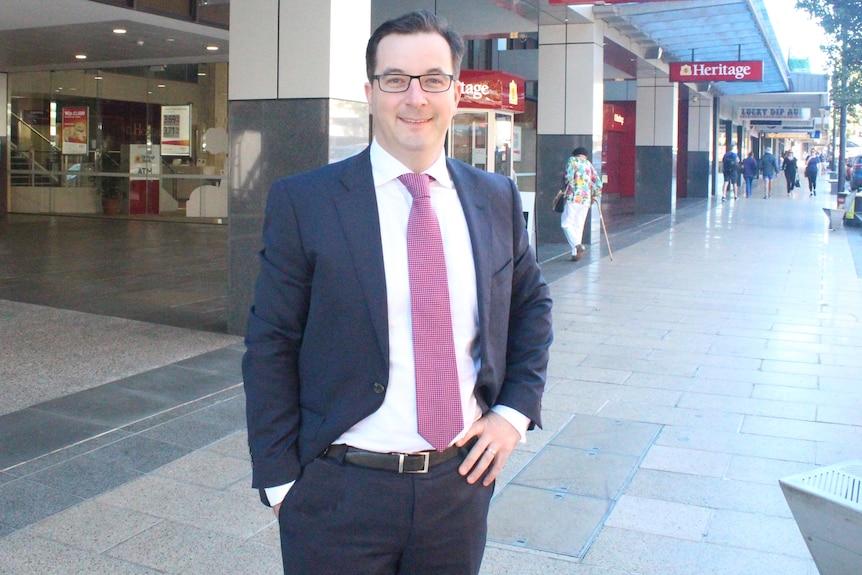 A middle-aged smiling man with glasses wearing a suit on the street, pink tie.