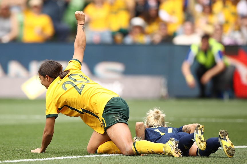 A female soccer player wearing yellow and green puts her fist in the air as she's on the ground