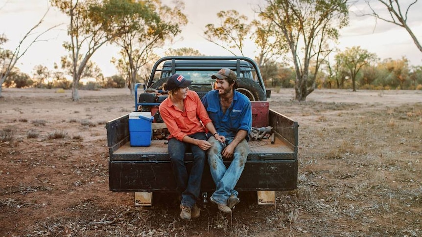 A woman and man sitting on the back of a ute in scrubland, with trees in the background.