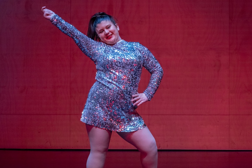 A dancer wearing a sparkly long sleeved dress poses with arm in the air against a red backdrop